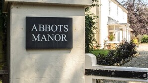 Un cartel con la leyenda "ABBOTS MANOR" en una pared blanca, con una entrada porticada y un edificio al fondo.