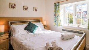 A neatly made bed with white linens and a teal pillow, a bedside lamp, and a window with floral curtains. - Poppy Cottage (Blandford Forum)