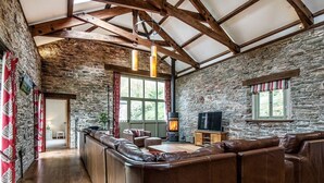 A rustic interior with a stone wall, wooden beams, a leather sofa, a fireplace, and a television. - Mill Barn (Kingsbridge)