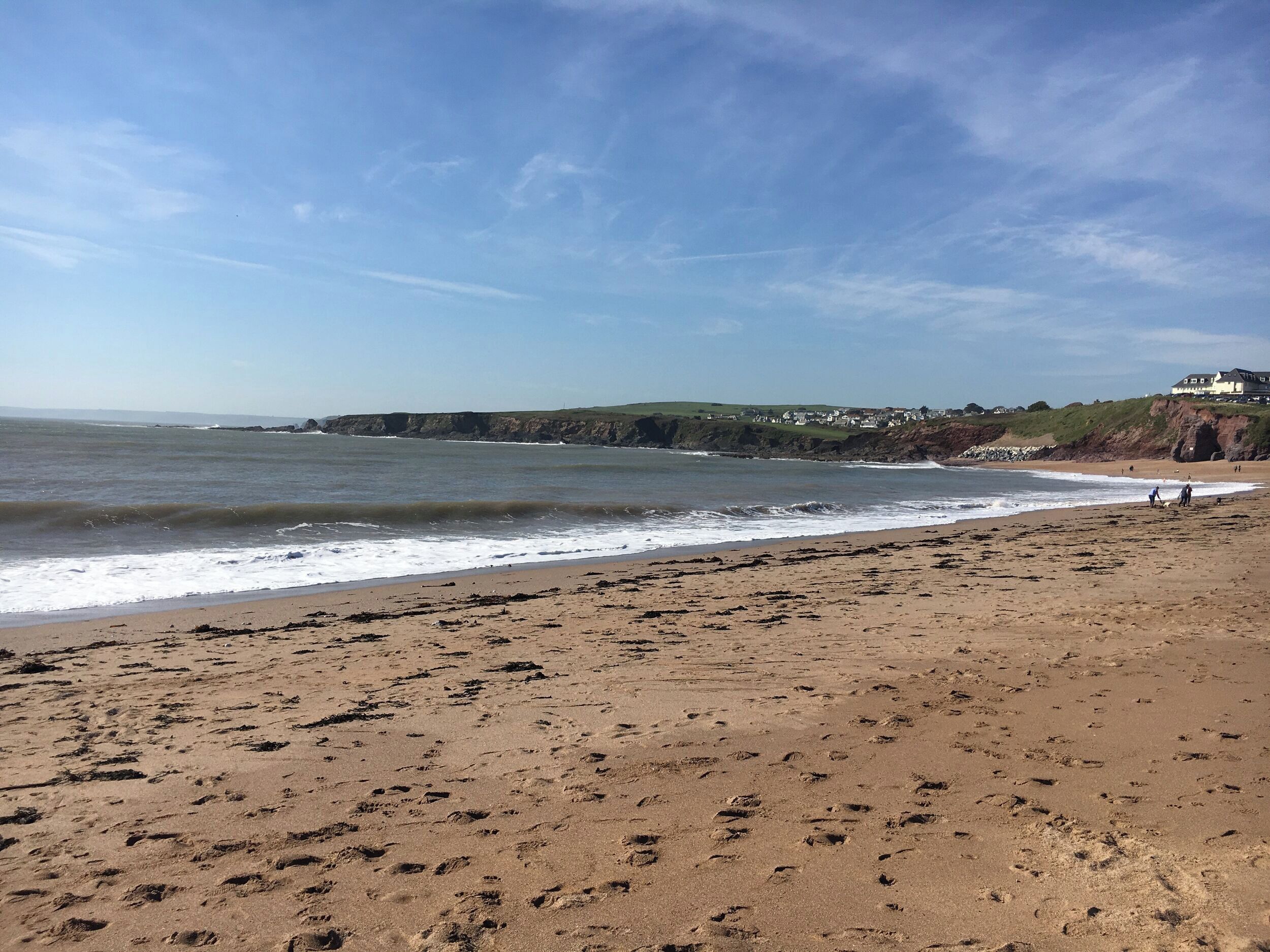 A sandy beach with footprints leading to the water, a clear sky, and a distant view of houses on a cliff.