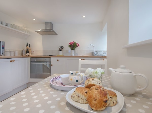 Una cocina moderna con gabinetes blancos, una campana extractora de acero inoxidable y un mantel de lunares sobre la encimera. Hay platos con scones y una tetera blanca sobre la mesa.