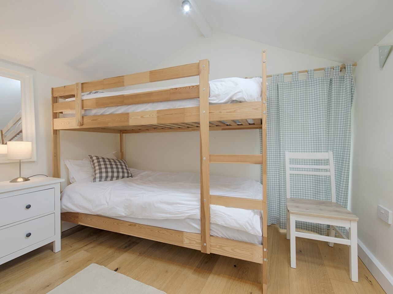 A bunk bed with a white mattress and a checkered pillow, a white nightstand with drawers, a white chair, and a checkered curtain.