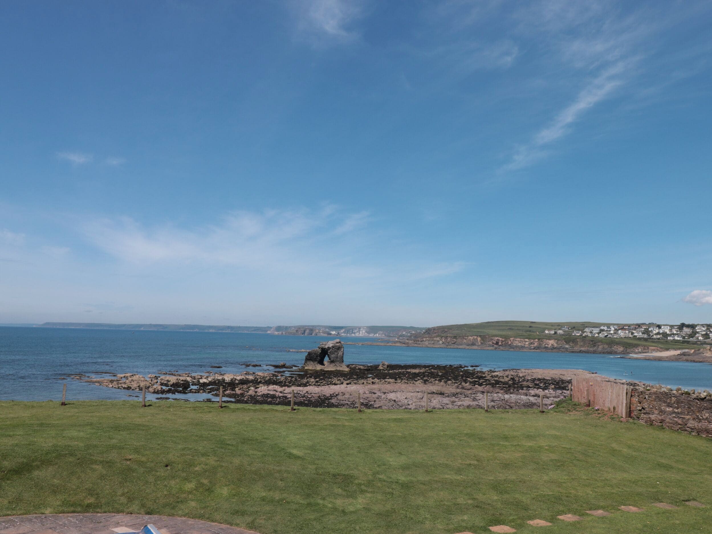 Un paysage côtier avec une zone herbeuse, une côte rocheuse et une vue lointaine sur des maisons.