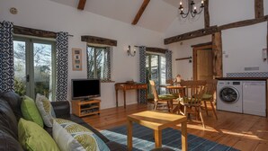 A cozy living room with a sofa, TV, wooden dining table, and chairs. There's a kitchen area with a washing machine and a dining table. - Bradbridge Barn (Plymouth)