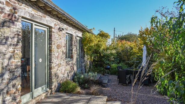 A stone building with a glass door, a patio with a grill, and a garden with various plants. - Bradbridge Barn (Plymouth)