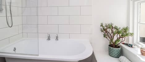 Une salle de bain avec une baignoire, une douche, des murs recouverts de carreaux blancs, une plante en pot et une fenêtre.