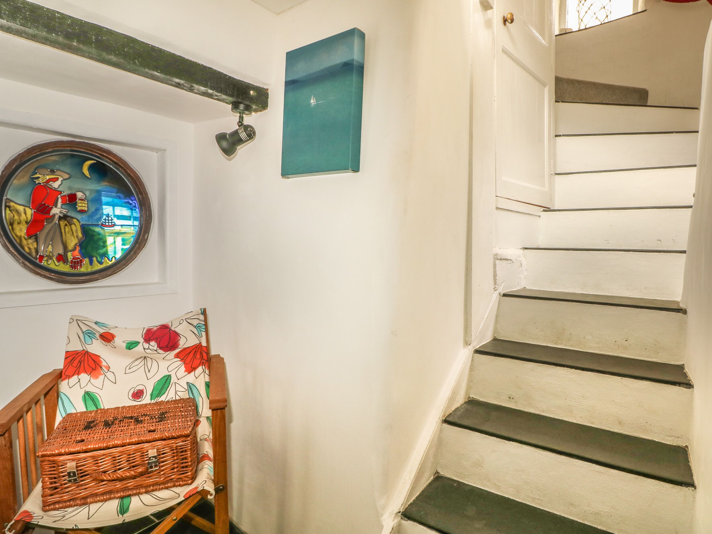 A staircase with white steps and a wooden handrail, a wall-mounted light fixture, a framed picture, and a wicker basket on a chair.