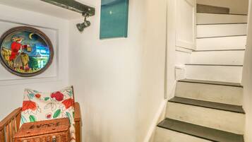 A staircase with white steps and a wooden handrail, a wall-mounted light fixture, a framed picture, and a wicker basket on a chair.