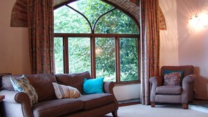 Uma sala de estar aconchegante com uma grande janela em arco, um sofá de couro marrom, uma poltrona combinando e uma almofada decorativa estampada.