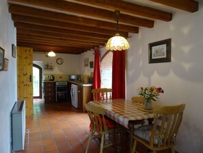 A cozy kitchen with wooden beams, a dining table with chairs, a hanging light fixture, and a clock on the wall.
