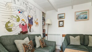 A cozy living room with two green sofas, patterned cushions, a wooden coffee table, a bookshelf, and a mural on the wall. - Mural Cottage (Penzance)