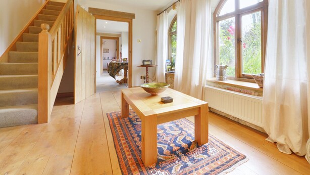 Een ruime kamer met een houten trap, een groot raam met gordijnen, een houten tafel met een kom en boeken, en een kamerkleed met een patroon.