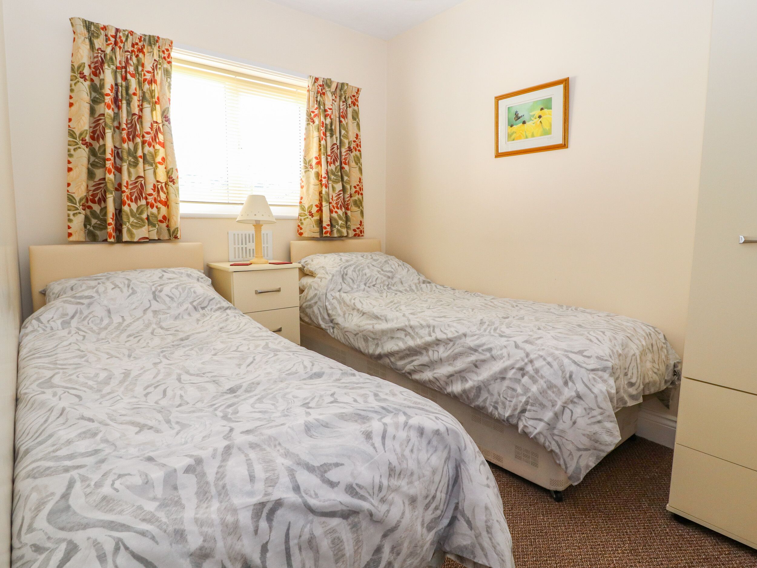 A bedroom with two beds, a nightstand, a lamp, a framed picture on the wall, and a window with curtains.