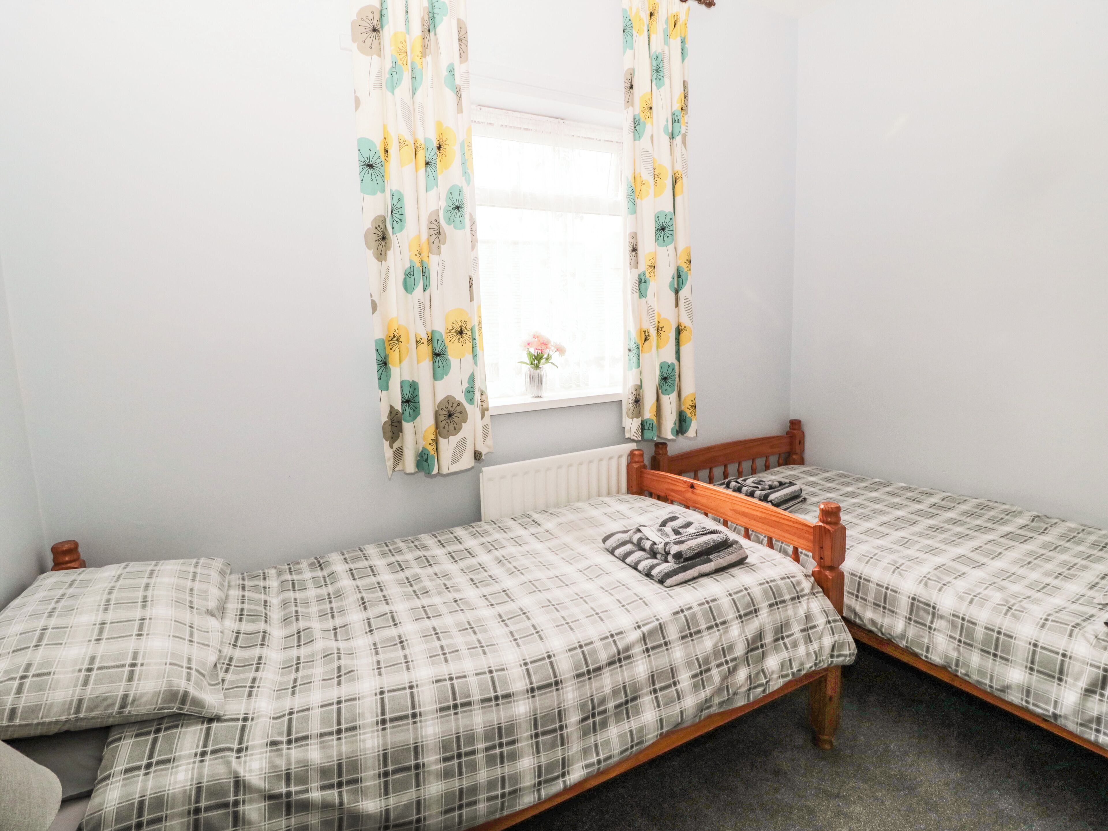 A room with two single beds, a window with curtains, a radiator, and a small table with a vase of flowers.
