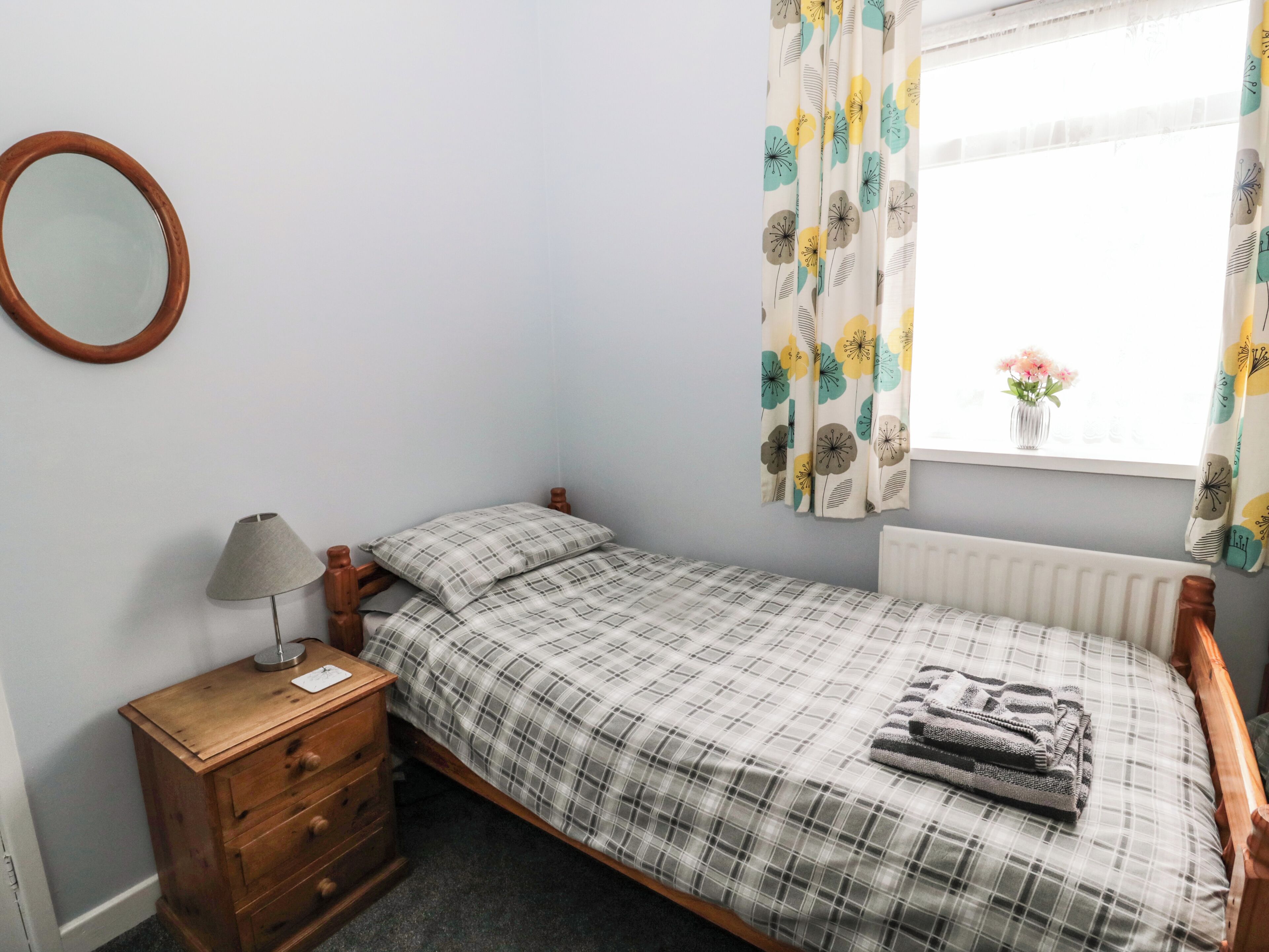 Een kleine slaapkamer met een enkel bed, een houten nachtkastje, een lamp, een dressoir en een raam met bloemen gordijnen.