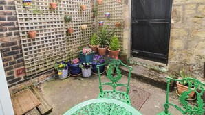 Une petite cour avec un mur de briques, une treille en bois et une porte noire. On y trouve plusieurs plantes en pot et une chaise en métal verte.