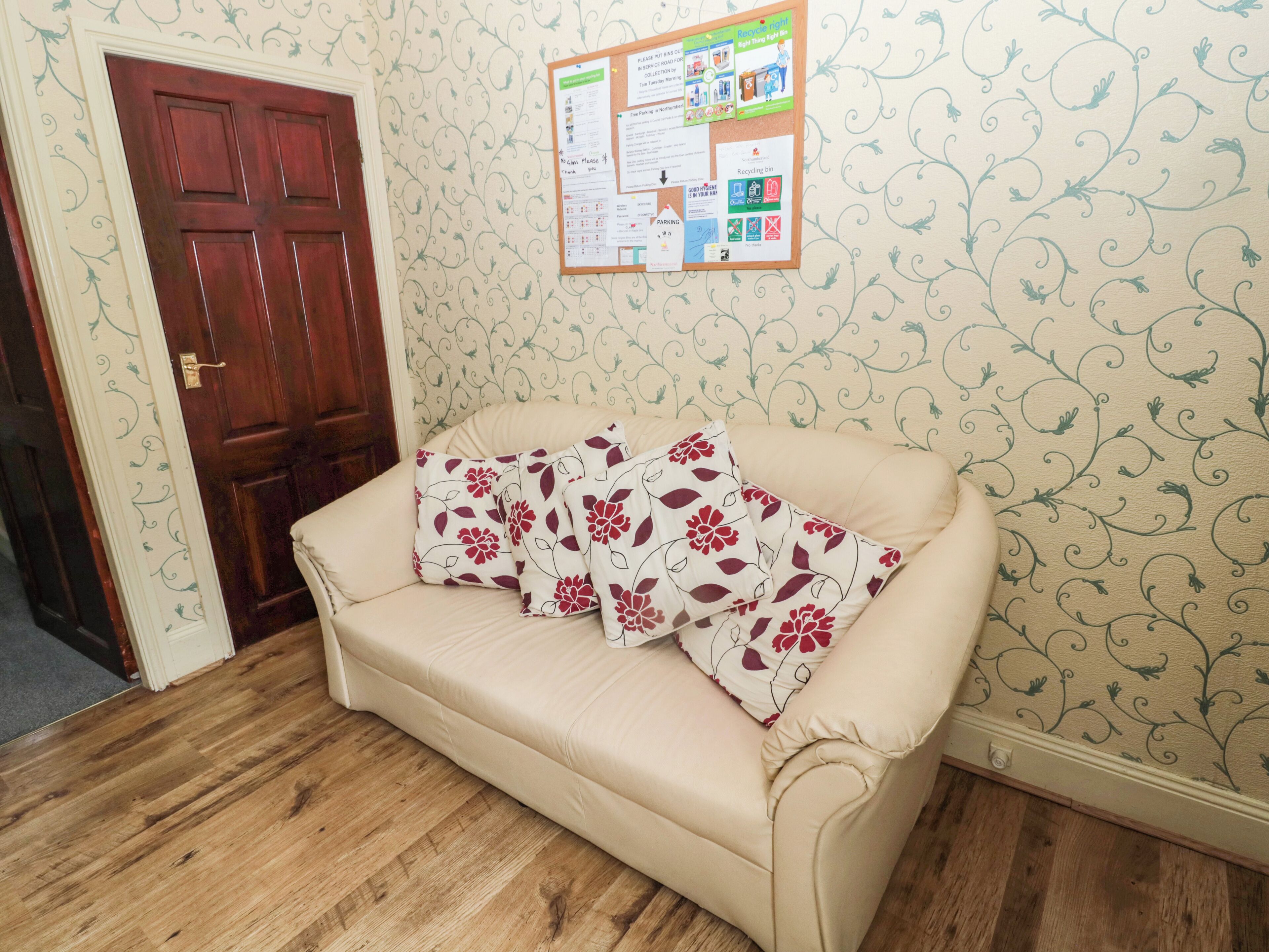 A room with a wooden floor, a beige sofa with floral cushions, a wooden door, and a bulletin board with various notices.