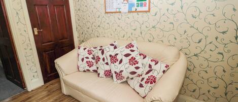 A room with a wooden floor, a beige sofa with floral cushions, a wooden door, and a bulletin board with various notices.