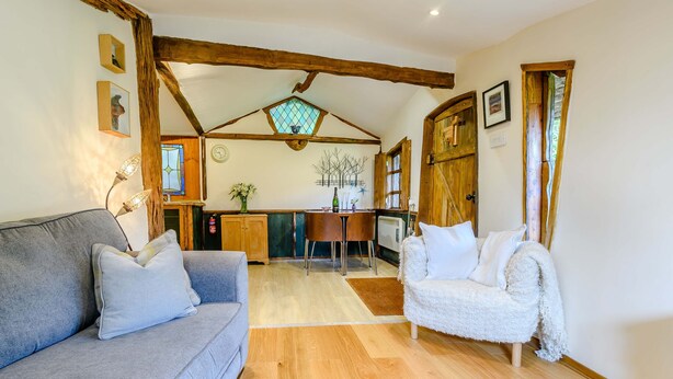 A cozy living room with a grey sofa, a white armchair, wooden beams, a dining area with a table and chairs, and a kitchen in the background. - Brookbank Folly (Mitcheldean)