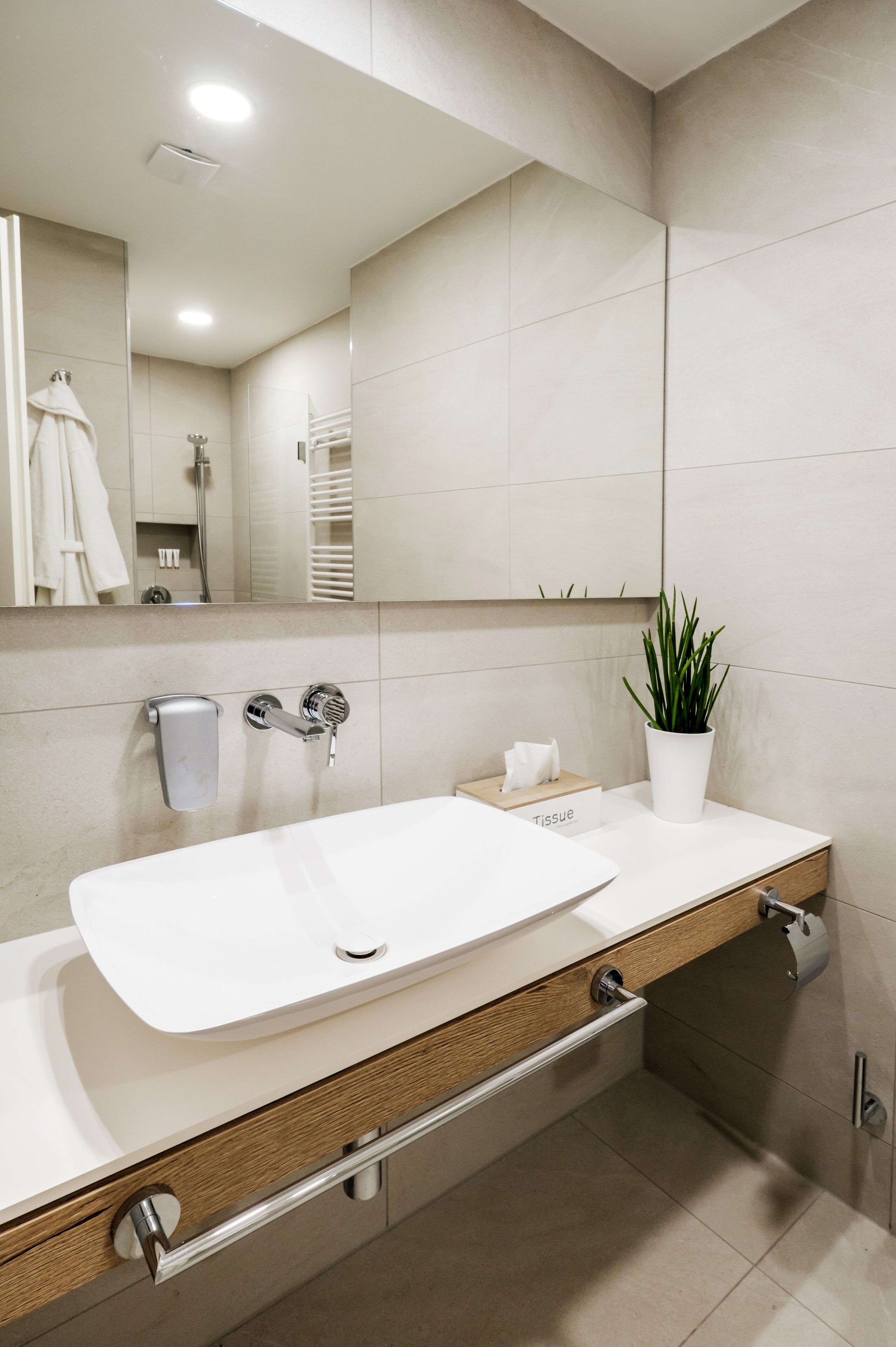 Un baño moderno con un lavamanos grande blanco, un espejo, un perchero para toallas, una planta y una caja de pañuelos.
