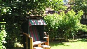 Una silla de madera al aire libre con un dosel estampado, ubicada en un jardín con abundante vegetación y una estructura de techo con tejas al fondo.