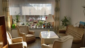 Una sala de estar acogedora con un sofá, sillones y una mesa de centro. Hay plantas en macetas en un alféizar, y una lámpara sobre una mesita auxiliar.
