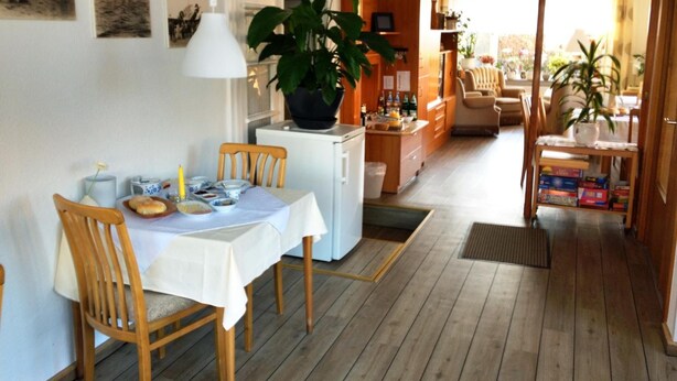 Un comedor con una mesa lista para dos, una planta en maceta y una cocina al fondo.