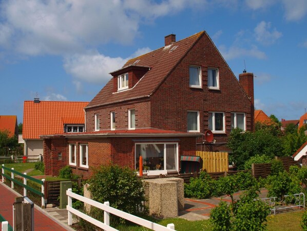 A two-story brick house with a red-tiled roof, a visible chimney, and a front yard with a wooden fence and greenery. - Pension Friedrich Voss (Langeoog)