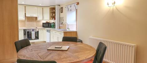 A dining area with a round wooden table and four chairs, a kitchen in the background with white cabinets and a microwave, and a white radiator on the wall.