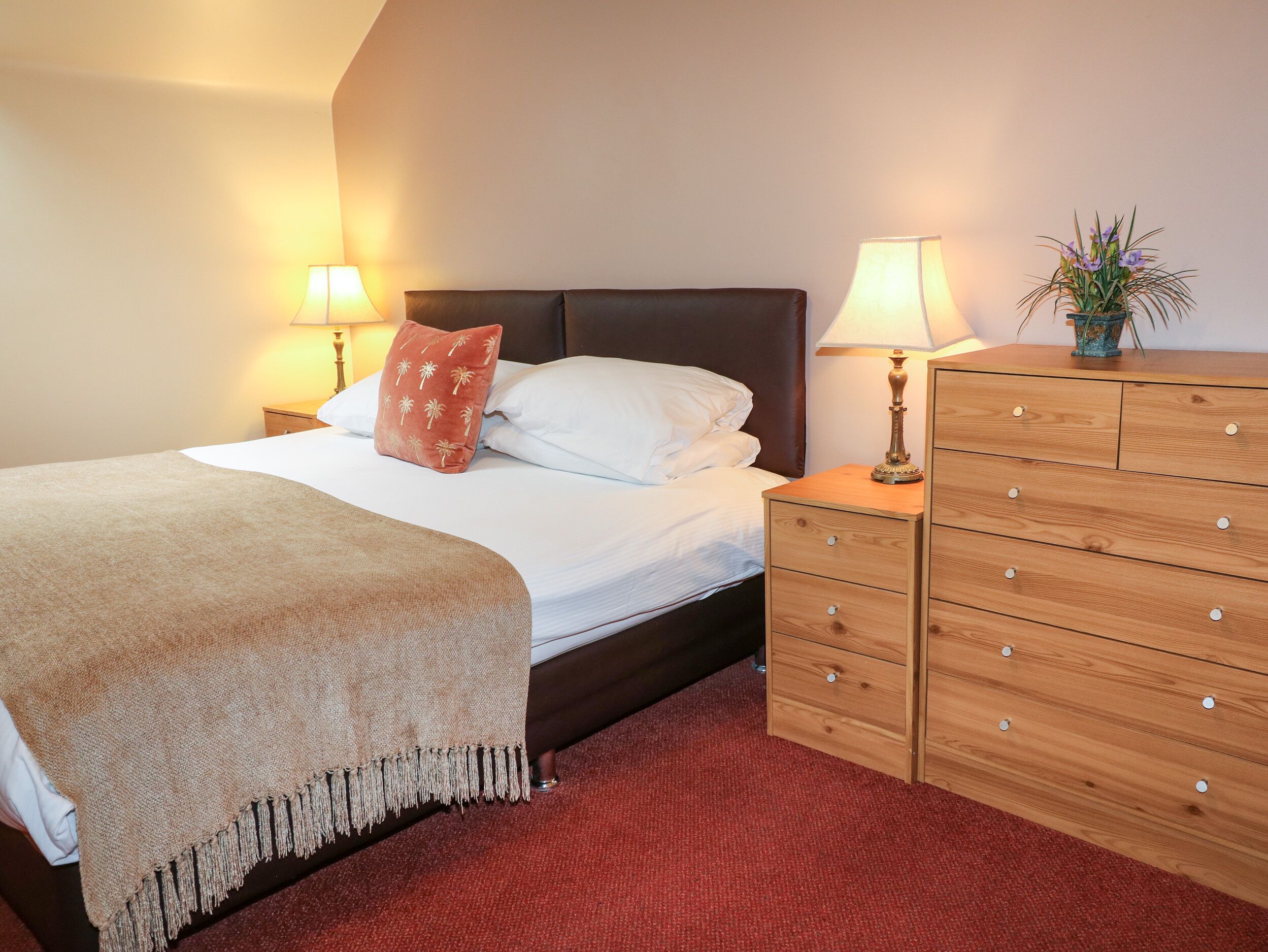 A neatly made bed with a brown throw blanket, a wooden dresser with drawers, and two bedside lamps.
