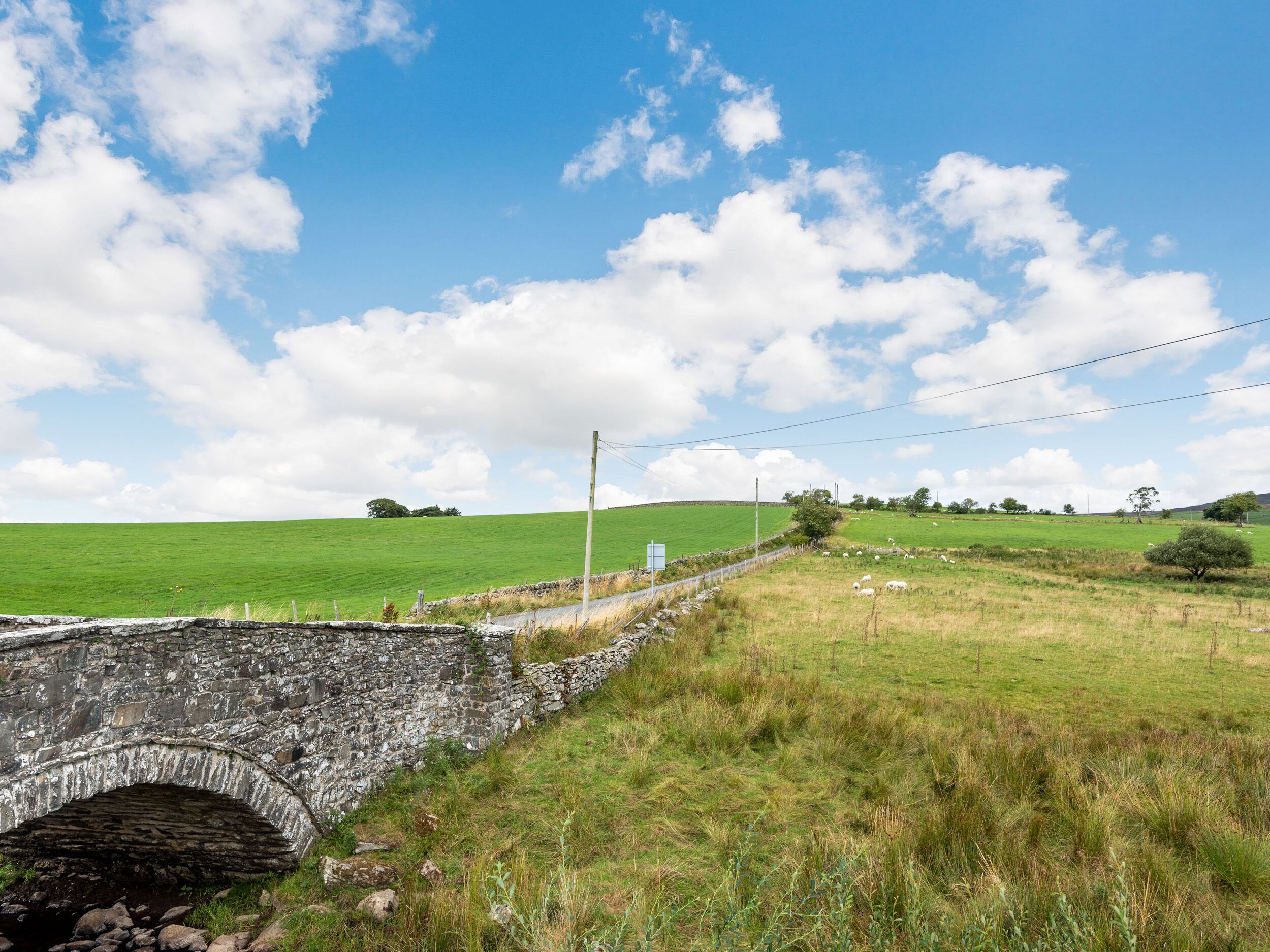A stone bridge over a stream, surrounded by green fields and a clear blue sky.
