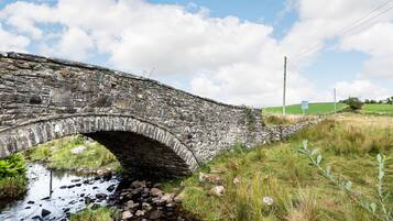 A stone bridge over a stream with a grassy field and a clear sky in the background.