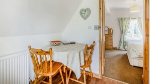 Een gezellige kamer met een houten eettafel, twee stoelen, een klok in de vorm van een hart, en een uitzicht op een slaapkamer door een open deur.