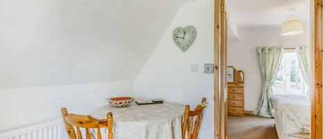 A cozy room with a wooden dining table, two chairs, a heart-shaped clock, and a view of a bedroom through an open door.