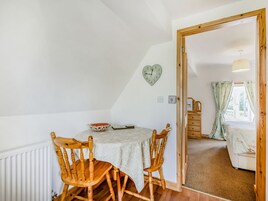 A cozy room with a wooden dining table, two chairs, a heart-shaped clock, and a view of a bedroom through an open door.