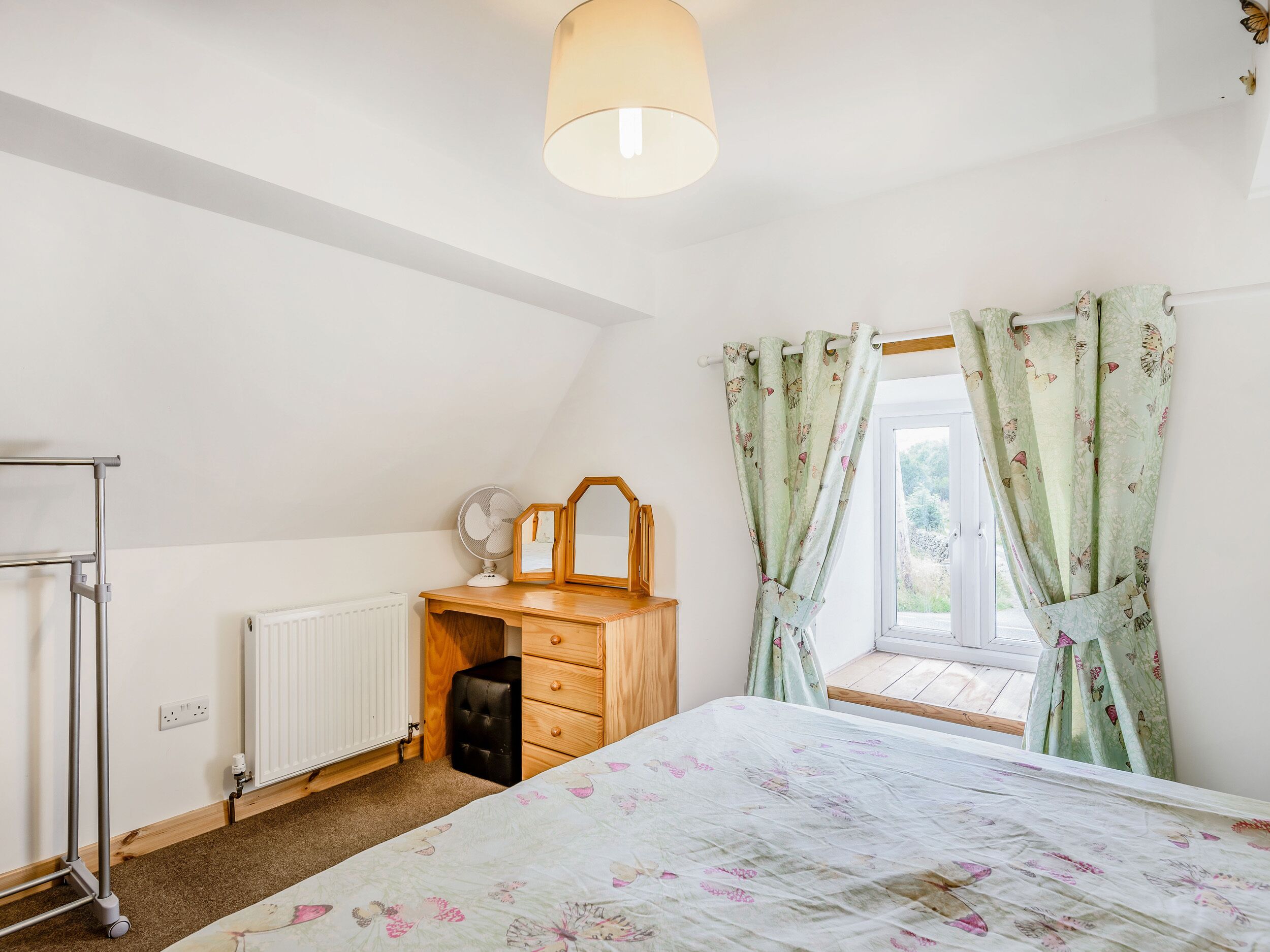 A bedroom with a bed, a wooden dresser, a mirror, a fan, a radiator, and a window with curtains.