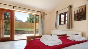 Un dormitorio con una cama grande, una manta roja y toallas sobre la cama. Hay una ventana con cortinas, un radiador y un cuadro en la pared.