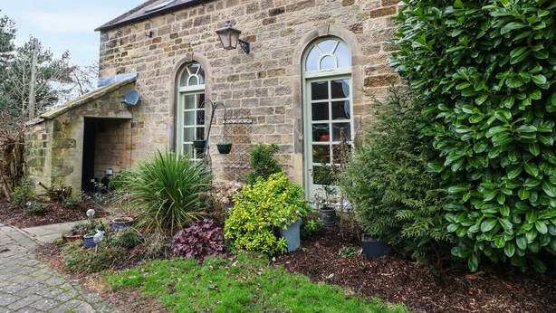 A stone building with two arched windows, a small door, and a garden with various plants. - The Old Chapel (Chesterfield)