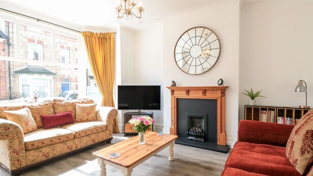 Un salón con sofá, un sillón rojo, una mesa de centro de madera, chimenea, televisión y un ventanal con cortinas.