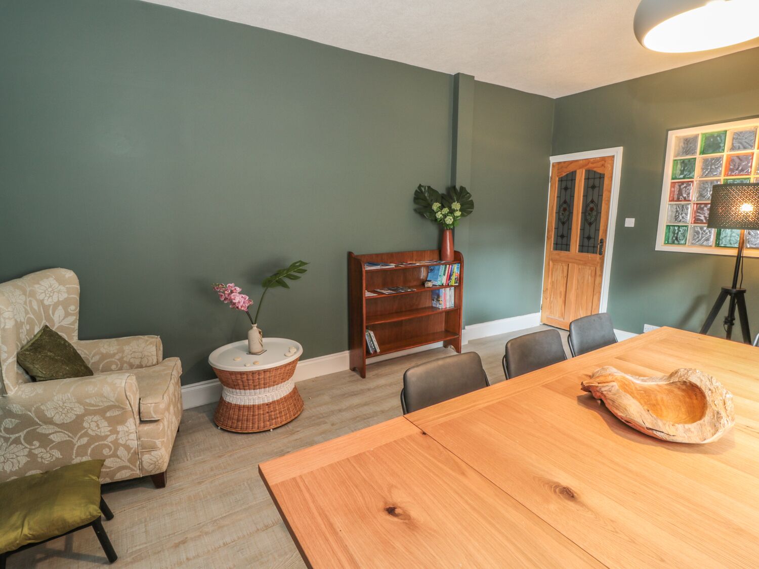A dining area with a wooden table and chairs, a bookshelf, a wicker side table with a vase of flowers, and a green wall.