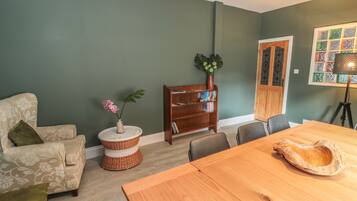 A dining area with a wooden table and chairs, a bookshelf, a wicker side table with a vase of flowers, and a green wall.