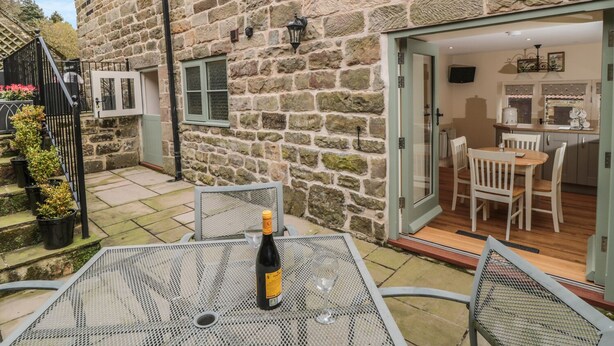 A stone building with a patio area featuring a table with a bottle and glass. - New Stable Cottage (Whitby)