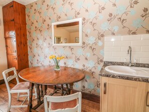 Een eethoek met een ronde houten tafel, twee witte stoelen, een vaas met gele bloemen, een houten kast en een spiegel aan de muur.