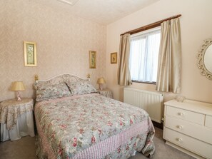 Een slaapkamer met een bloemenbedekking, twee nachtkastjes met lampen, een kaptafel, een raam met gordijnen en een radiator.