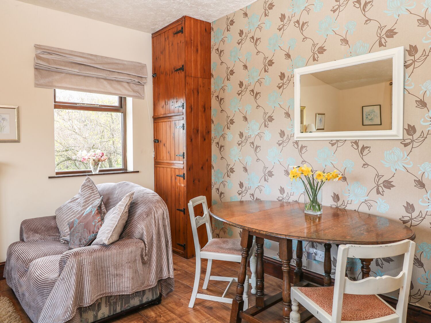Une pièce chaleureuse avec une armoire en bois, un papier peint à motifs floraux, une table de salle à manger ronde en bois entourée de chaises, et une fenêtre donnant sur les arbres.