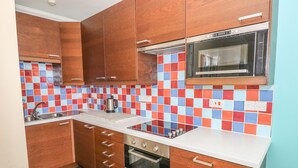 A compact kitchen with wooden cabinets, a microwave, a stove, and a tiled backsplash. - Apartment 2 (Tenby)