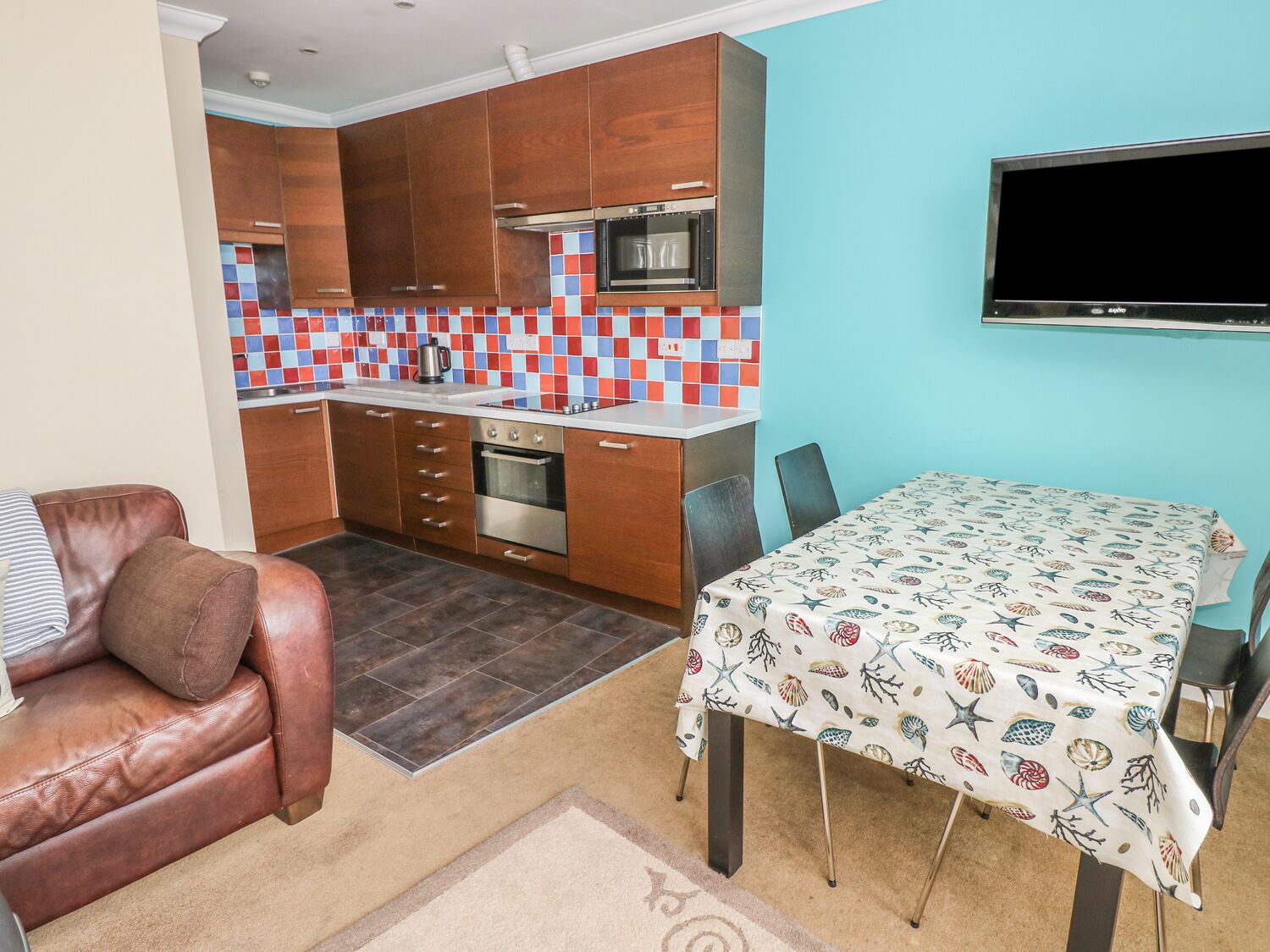 Una cocina compacta con armarios de madera, salpicadero de azulejos y horno empotrado. Hay una zona de comedor con una mesa cubierta con un mantel estampado con motivos de conchas.