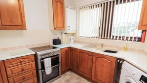 A kitchen with wooden cabinets, a stove, a washing machine, and a window with blinds. - Harbour Side (Maryport)