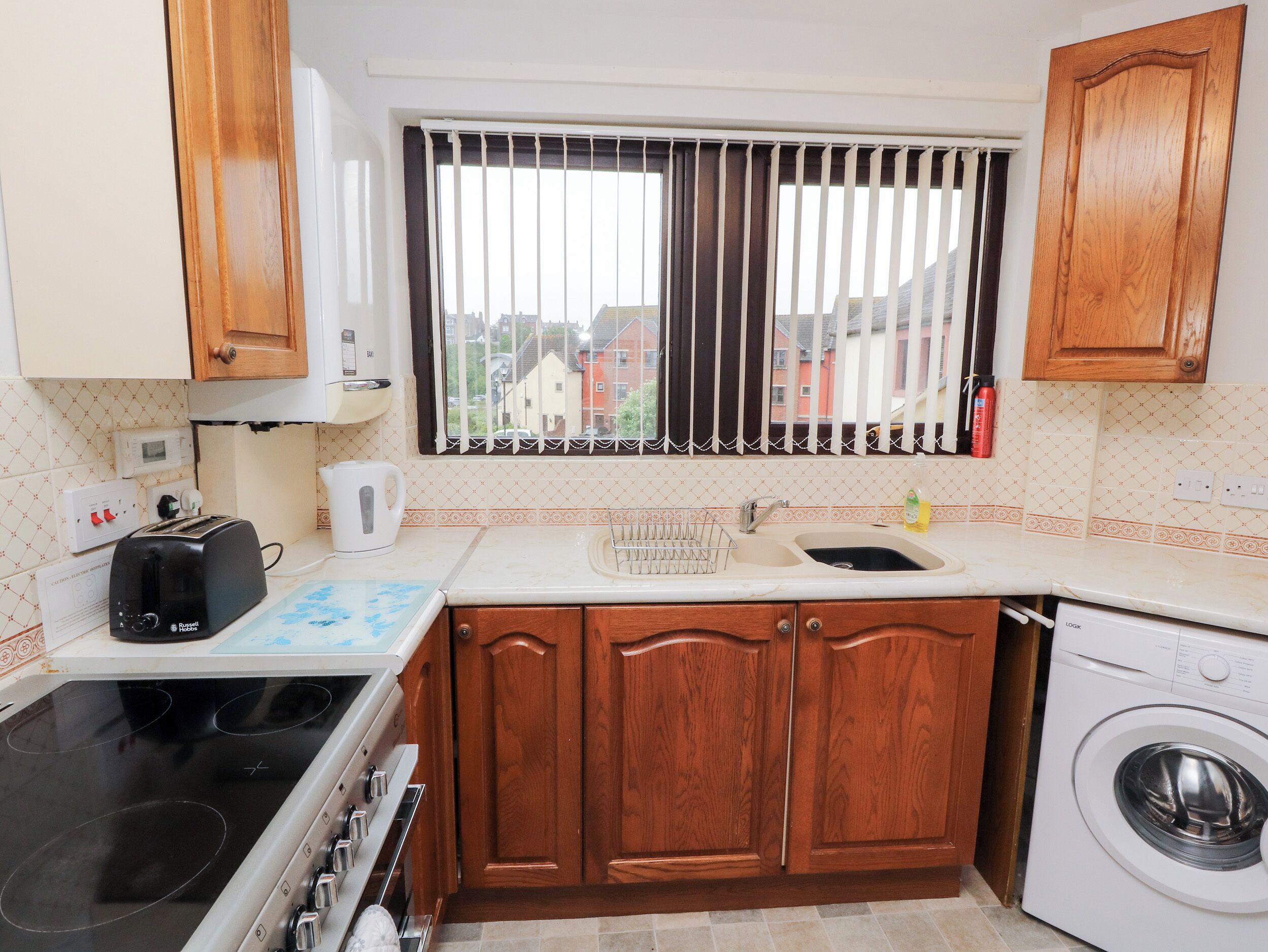 A kitchen with wooden cabinets, a stove, a washing machine, a window with blinds, and a view of buildings outside.
