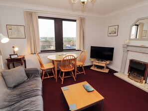 A furnished room with a sofa, a dining table with chairs, a television, a fireplace, and a window with curtains. - Harbour Side (Maryport)